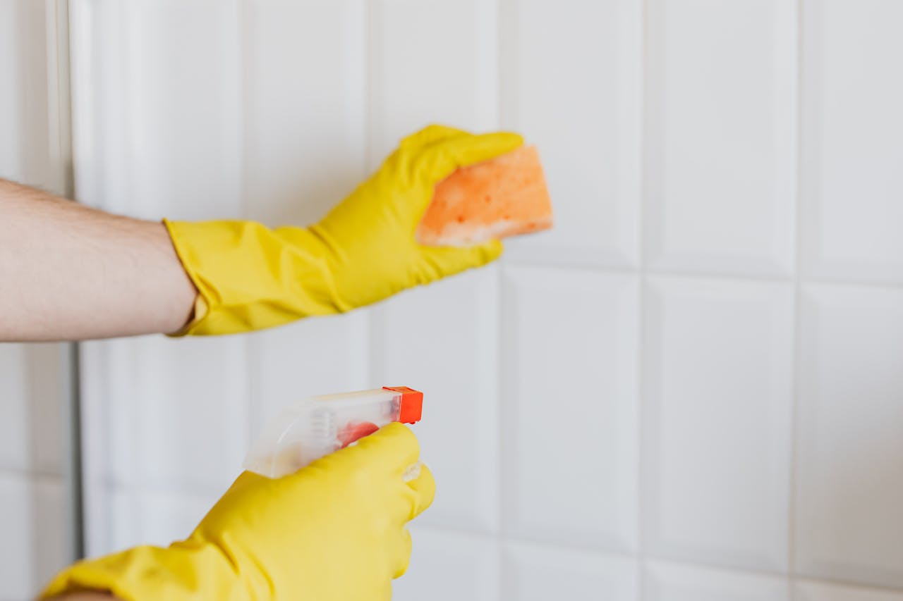 hero-01 A person wearing yellow gloves cleans bathroom tiles with a sponge and spray bottle.